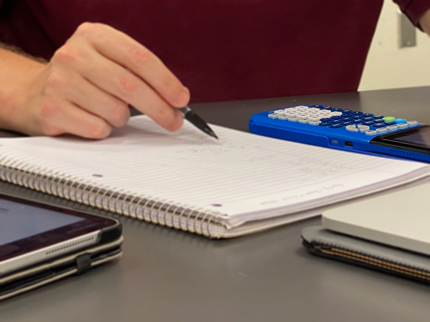 A student's hand writing in a notebook with a calculator an iPad next to it
