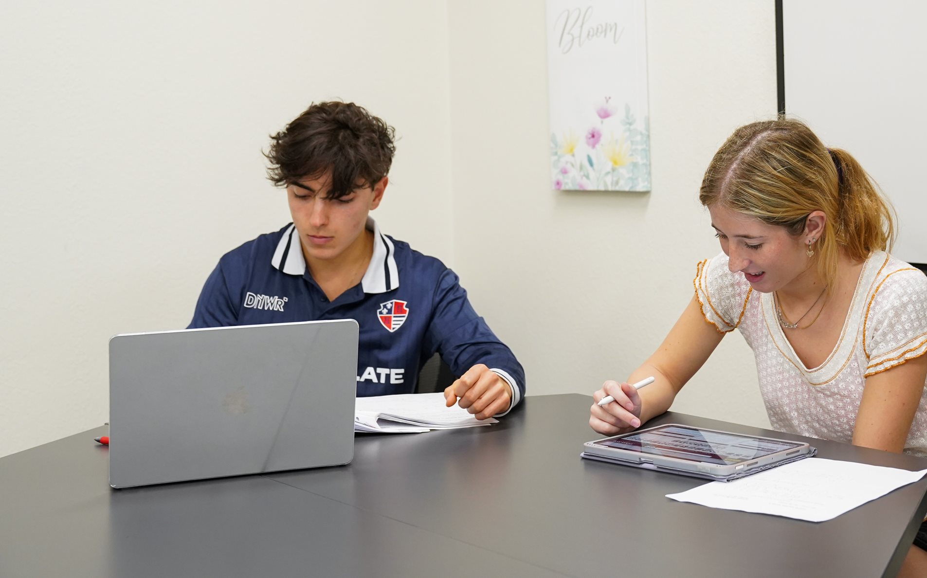 Two students working at a table, one of them writing on an iPad and the other using a computer