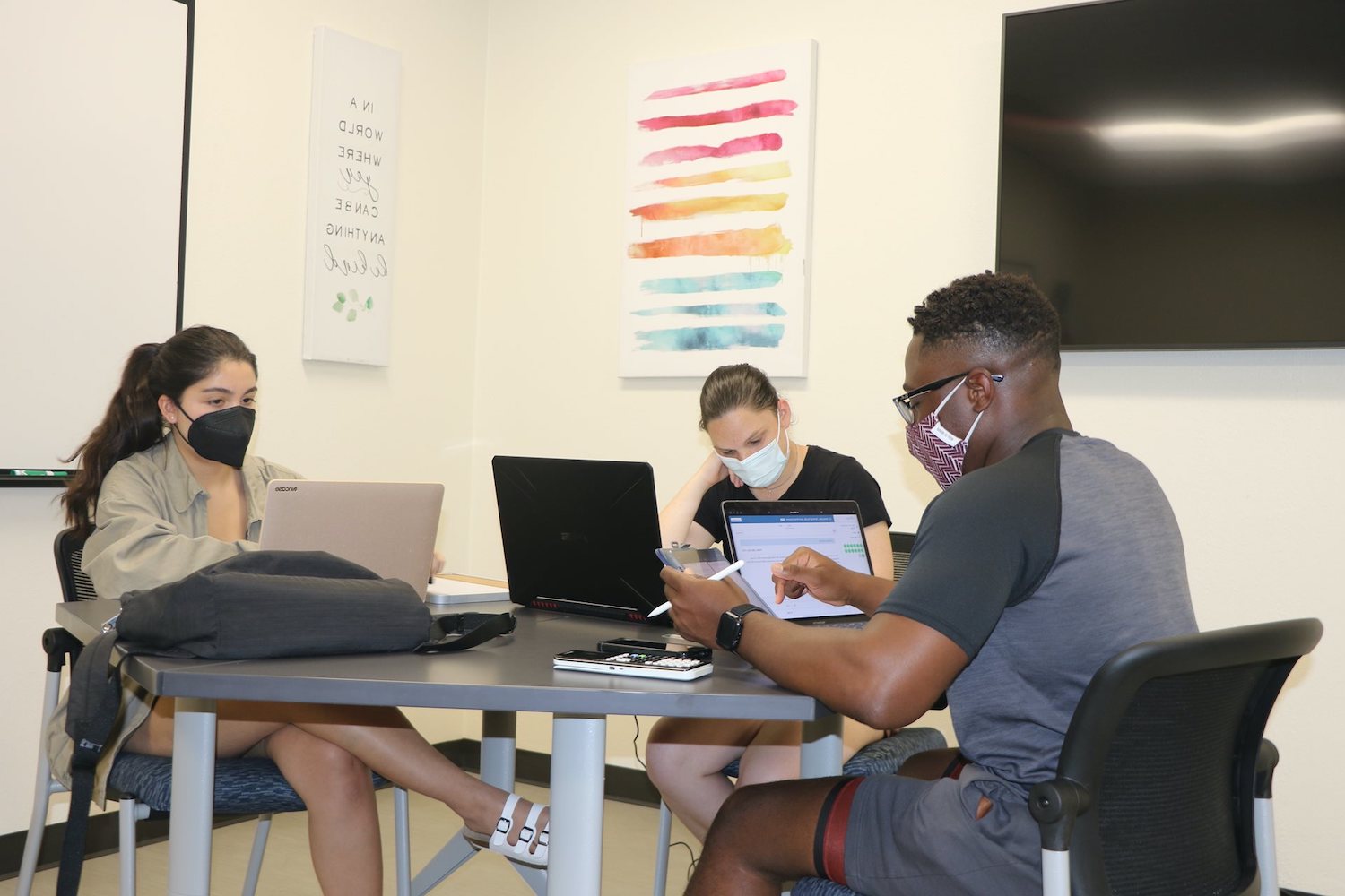 A group of three students working on laptops at a table, one also has an iPad and calculator